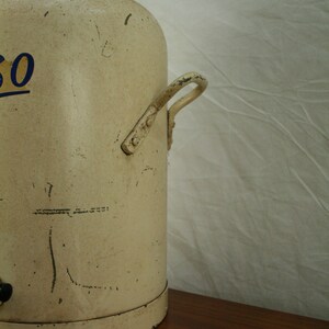 May include: A vintage white metal water cooler with a silver lid and a metal handle. The cooler has a blue and yellow inscription on the side.