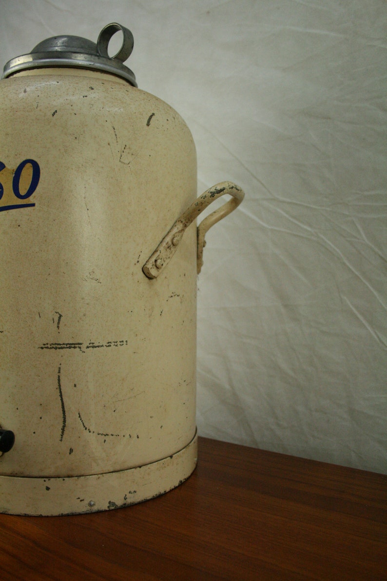 May include: A vintage white metal water cooler with a silver lid and a metal handle. The cooler has a blue and yellow inscription on the side.