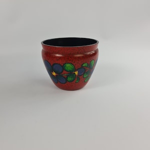 May include: A red ceramic planter with a floral design featuring blue flowers and green leaves. The planter has a black rim.