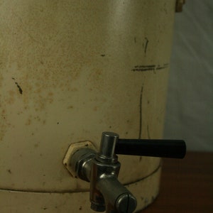 May include: A white metal water cooler with a silver spigot and a black handle. The cooler has a worn, aged appearance.