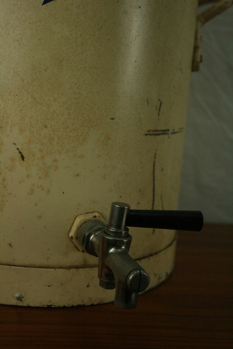 May include: A white metal water cooler with a silver spigot and a black handle. The cooler has a worn, aged appearance.