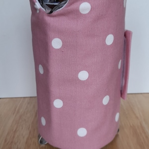 May include: A pink French press cosy with white polka dots. The cosy is made of fabric and fits snugly around the stainless steel French press. The French press has a black knob on top and sits on small metal feet.