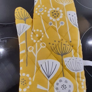 May include: Mustard yellow oven glove with a floral design in white and grey. The quilted glove has a white trim and features stylized flowers and seed heads. Designed to protect hands from heat when handling hot items.