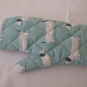 May include: A quilted oven glove featuring a light teal background with a repeating pattern of white dachshund dogs with black accents. It has a black trim and a white hanging loop.