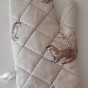 May include: Beige quilted oven glove with a brown otter print. The glove has a diamond pattern and a brown trim. A white loop is attached for hanging. The otter design includes details of grass and reeds.
