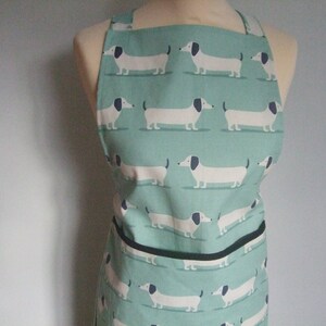 May include: A teal apron with a repeating pattern of white dachshund dogs with black ears and tails. The apron has a black pocket and trim.