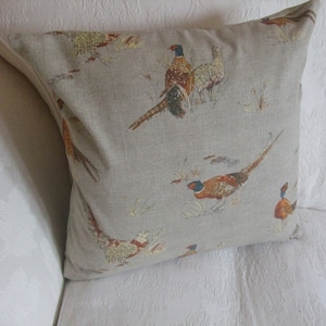 May include: Square cushion featuring a pheasant design on a beige background. The birds are depicted in brown, orange, and blue. The cushion is placed on a white surface.