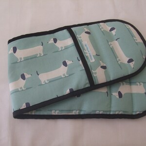 May include: Oven gloves featuring a light teal background with a repeating pattern of white dachshund dogs with black ears and tails. The gloves are edged with black binding.