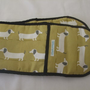 May include: An olive green double oven glove featuring a repeating pattern of white and grey dachshund dogs. The edges are trimmed with black binding. A small label with text is visible. The item is designed for kitchen use.