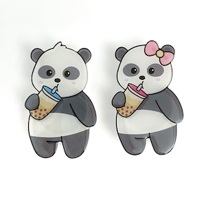 May include: Two panda bear cartoon character charms with black and white fur, holding bubble tea with pink and blue straws. The panda on the left has no accessories. The panda on the right has a pink bow on its head.