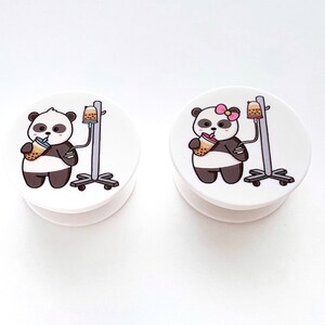 May include: Two white phone grips with a panda bear design. The panda bears are wearing a pink bow and holding a boba tea. The phone grips are attached to a clear plastic stand.