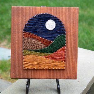 May include: A wooden square with a string art design of a night sky with a full moon and hills. The string art is done in shades of blue, green, orange, and brown.