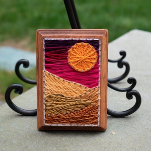May include: A framed string art piece depicting a sunset with a large orange sun and pink and orange mountains. The string art is in a wooden frame.