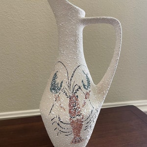 May include: White ceramic pitcher with a textured surface and a lobster design. The lobster is brown, red, and green.