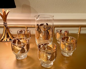 May include: A set of six clear glass drinking glasses with gold pear and fruit designs. The glasses are arranged on a gold surface. A matching pitcher is in the center of the set.