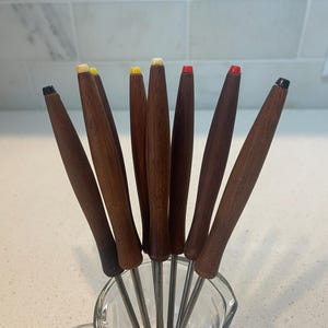 May include: A set of eight fondue forks with long, tapered dark brown wooden handles. Each handle is topped with a small, distinctively colored plastic cap in shades of red, yellow, white, or black, attached to thin, silver-toned metal shafts.