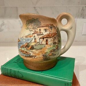 May include: A ceramic pitcher with a painted coastal scene featuring buildings, trees, and boats. The pitcher has a tan base and handle, with a detailed illustration in shades of brown, blue, and green. The pitcher is sitting on a green book.