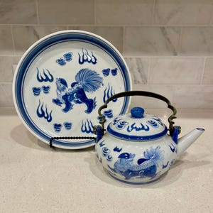 May include: A white and blue porcelain tea set featuring a plate and a teapot. The plate and teapot are decorated with blue illustrations of mythical creatures and floral patterns. The teapot has a metal handle and spout.