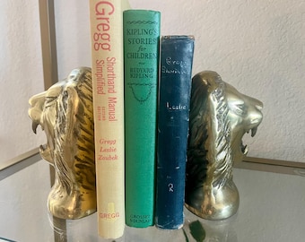 May include: A pair of brass lion head bookends holding three vintage books. The books are in shades of green, blue, and cream, with titles including "Kipling's Stories for Children" and "Gregg Shorthand Manual". The bookends add a decorative touch.