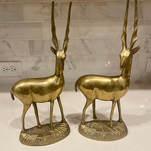 May include: A pair of gold-toned brass gazelle statues. The gazelles are standing on a base with a textured surface. The statues are about 10 inches tall.