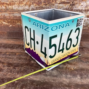 May include: Square planter made from an Arizona license plate. The planter features the license plate number CH-45463, with the words "ARIZONA" and "GRAND CANYON STATE" visible. The planter has a metal interior and is riveted together.