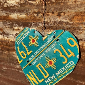 May include: A heart-shaped decoration made from a New Mexico license plate. The teal plate features yellow lettering and a red and yellow sun symbol. The text includes "New Mexico" and "Land of Enchantment."