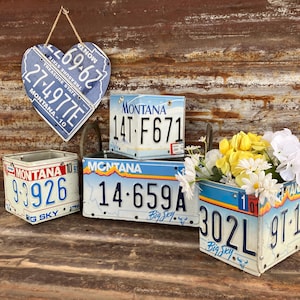 May include: A collection of decorative planters and a heart-shaped wall hanging, all crafted from repurposed Montana license plates. The planters are filled with yellow and white flowers. The heart-shaped piece is suspended by a twine string.