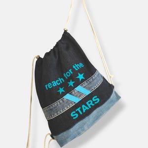 Op de afbeelding: Een zwarte trekkoordtas met een denim ontwerp. De tas heeft de tekst "reach for the stars" en drie blauwe sterren erop gedrukt.