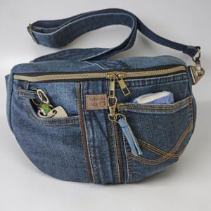 Könnte beinhalten: Eine blaue Bauchtasche aus Jeansstoff mit verstellbarem Riemen, einem Hauptfach mit Reißverschluss und mehreren Außentaschen. Ein brauner Aufnäher in Lederoptik trägt den Text Schicki Bag. Eine Tasche enthält Schlüssel, eine andere ein Objekt.