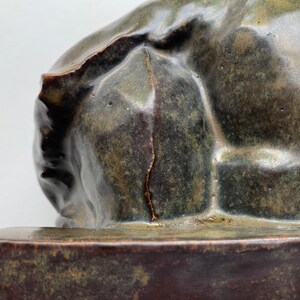 May include: A ceramic sculpture of a reclining figure with a rough, textured surface. The sculpture is a dark green color with brown and gold accents. The sculpture is sitting on a dark brown base.