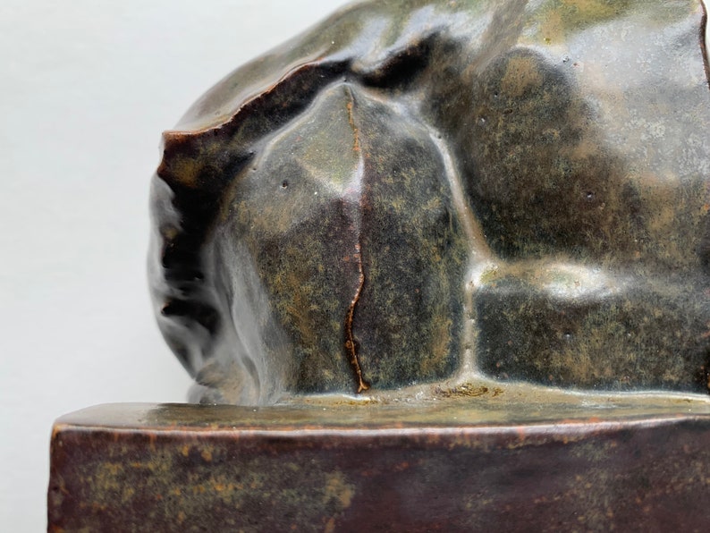 May include: A ceramic sculpture of a reclining figure with a rough, textured surface. The sculpture is a dark green color with brown and gold accents. The sculpture is sitting on a dark brown base.