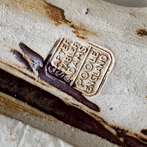 May include: A close-up of a ceramic object with a brown and white glaze. The object is stamped with the text 'Carter Stabler Adams Poole England'.