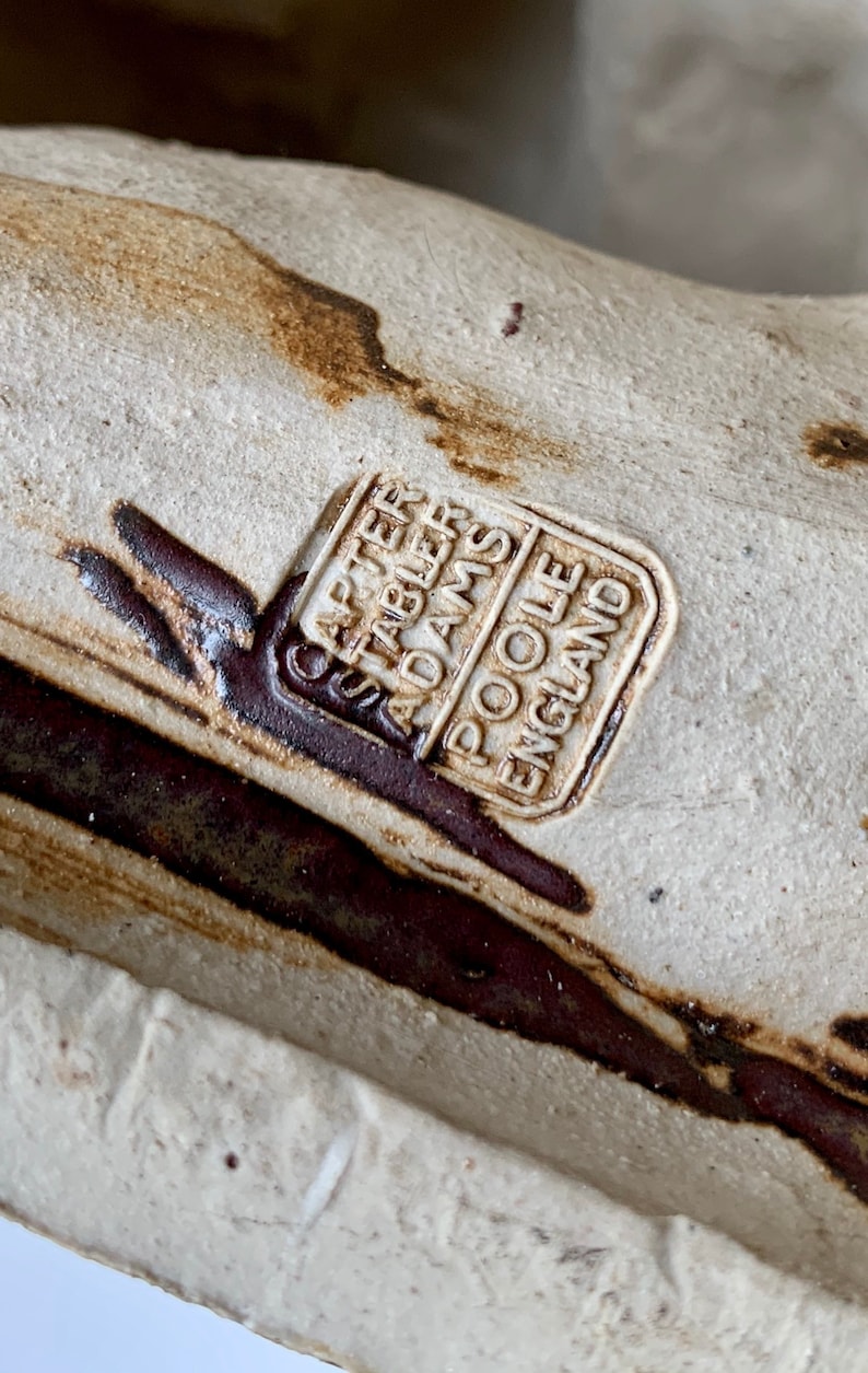May include: A close-up of a ceramic object with a brown and white glaze. The object is stamped with the text 'Carter Stabler Adams Poole England'.