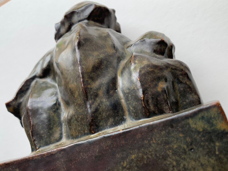 May include: A ceramic sculpture of two figures, one seated and the other standing, both with a dark green glaze. The sculpture is on a rectangular base.