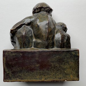 May include: A dark green ceramic sculpture of a person sitting with another person on their lap. The sculpture is on a dark green rectangular base.
