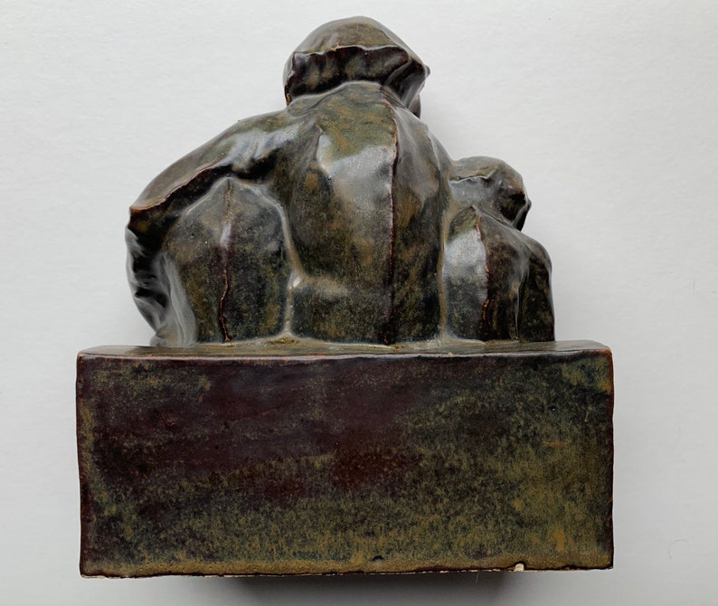May include: A dark green ceramic sculpture of a person sitting with another person on their lap. The sculpture is on a dark green rectangular base.