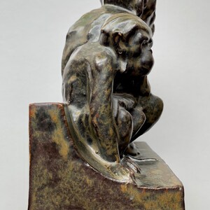 May include: A ceramic sculpture of two monkeys sitting on a ledge. The sculpture is green and brown with a speckled glaze. The monkeys are positioned with their backs to each other, and their heads are turned to look at each other.
