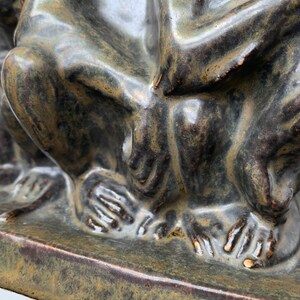 May include: Close-up of a ceramic sculpture of three figures sitting together. The sculpture is a dark green color with brown and gold accents. The figures are sitting on a square base.