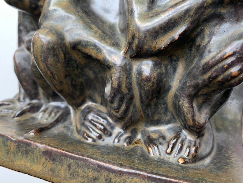 May include: Close-up of a ceramic sculpture of three figures sitting together. The sculpture is a dark green color with brown and gold accents. The figures are sitting on a square base.