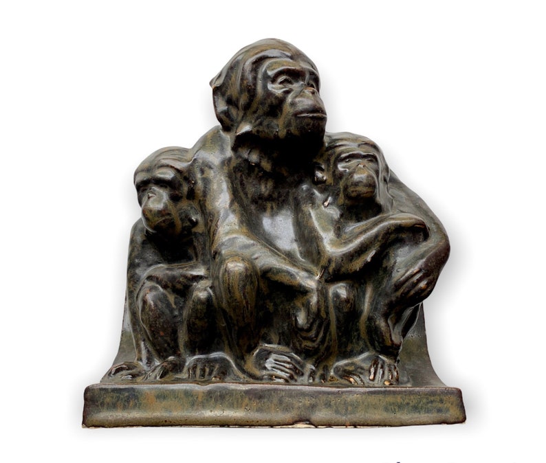 May include: A ceramic sculpture of three monkeys sitting together. The monkeys are all in a seated position, with the largest monkey in the center and the two smaller monkeys on either side. The monkeys are all made of a dark green ceramic and have a textured surface. The sculpture is on a rectangular base.