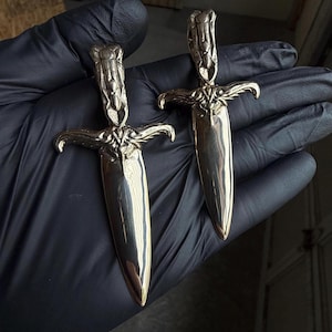 Pode incluir: Duas adagas prateadas ornamentadas, com cabos detalhados e guardas curvas. As lâminas são polidas e refletem a luz. As adagas são exibidas sobre um fundo escuro, destacando seu design e artesanato intrincados. Estes são provavelmente itens decorativos ou colecionáveis.