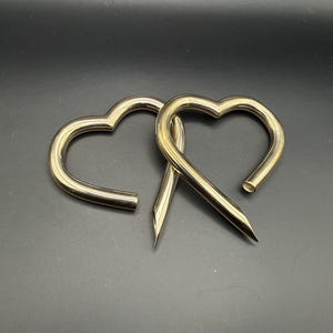 May include: Two metallic heart-shaped body jewelry pieces with a smooth, polished gold-tone finish. Each piece features a curved, open heart design with one end tapering to a sharp, pointed tip and the other end featuring a flat, circular opening.