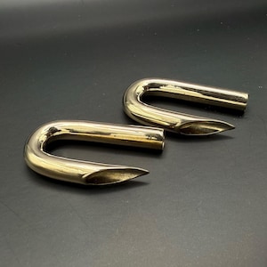 May include: Two polished, U-shaped metal objects with a golden hue. Each piece has a curved top and a tapered, pointed end. The objects are positioned on a dark surface, creating a contrast that highlights their form and shine.