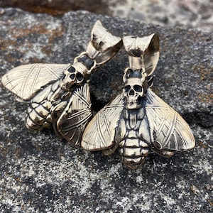 May include: Two gold-toned metal pendants shaped like moths with detailed wings and a skull head. The pendants have a textured surface and are hanging from a chain.