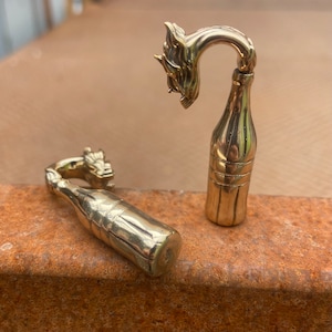 May include: Two gold bottle stopper ornaments shaped like a bottle of soda with a dragon head on the top. The bottle stoppers are on a rusty brown surface.