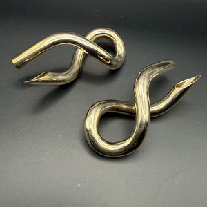 May include: Two polished gold-tone metal objects with a continuous, fluid, and twisted loop design. Each piece features a smooth, reflective surface that curves into an infinity-like shape with one end tapering to a pointed, open-ended tip.