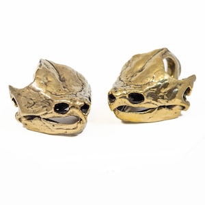 May include: Two brass-colored, stylized skull rings. The rings are detailed with textured surfaces and eye sockets. The rings are displayed on a white background. The rings are likely for jewelry or costume.