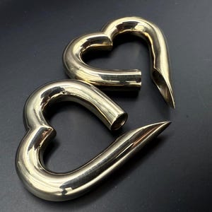 May include: Two polished brass heart-shaped objects. Each heart has a curved design with a pointed end. The objects are displayed on a dark surface, reflecting light.