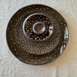 May include: A set of three nested circular ceramic dishes in a dark brown glaze with a hand-painted white polka dot pattern. The dishes are arranged concentrically, featuring a large plate, a medium bowl, and a small central bowl.