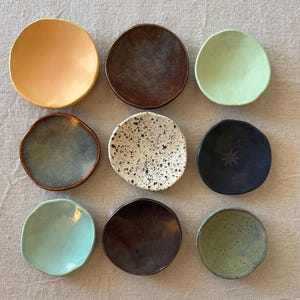 May include: A collection of nine small, round ceramic dishes arranged in a three-by-three grid. The dishes feature various finishes, including solid pastel yellow, dark brown, light green, blue-grey, white with black speckles, matte black with a star icon, light turquoise, deep brown, and olive green with dark flecks.